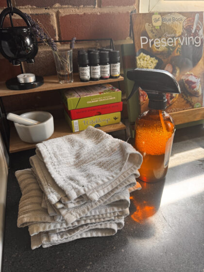 Amber spray bottle sitting on black counter next to white cleaning cloths. Behind that is a little shelf with a mortar and pestle, oracle card boxes, essential oil bottles, dried lavender sprigs in a shot glass, and a cauldron wax warmer. A book about canning is also seen in the background.