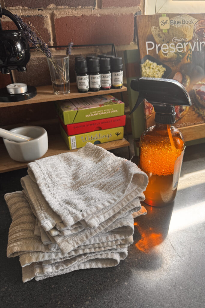 Amber spray bottle sitting on black counter next to white cleaning cloths. Behind that is a little shelf with a mortar and pestle, oracle card boxes, essential oil bottles, dried lavender sprigs in a shot glass, and a cauldron wax warmer. A book about canning is also seen in the background.