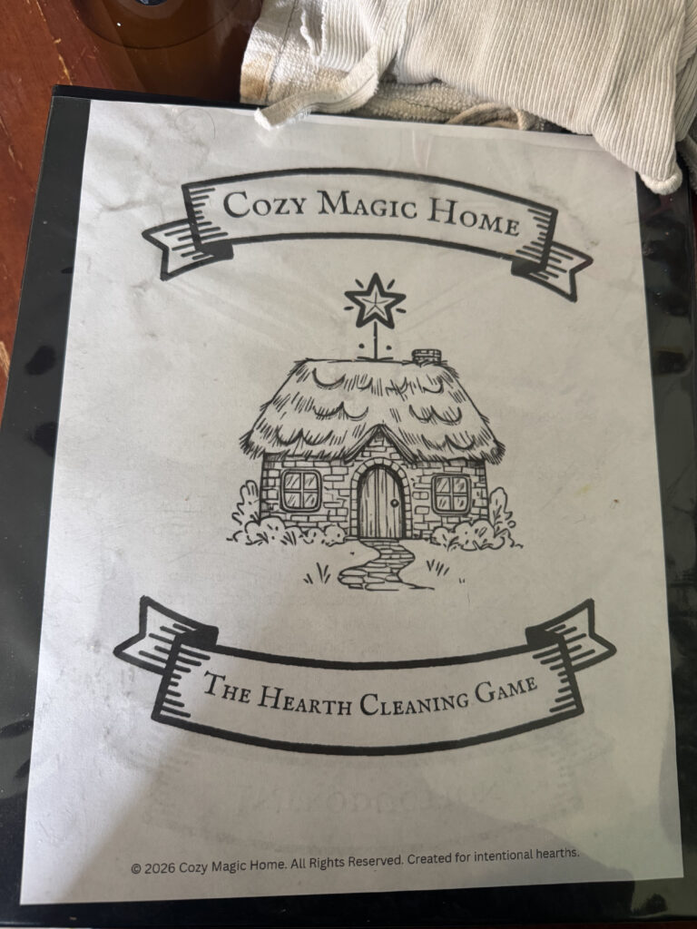 Black binder on a wooden table, and white cleaning cloths in one corner. Binder has a printed cover that says 'Cozy Magic Home', 'The Hearth Cleaning Game' and has the Cozy Magic Home logo in the middle in black and white