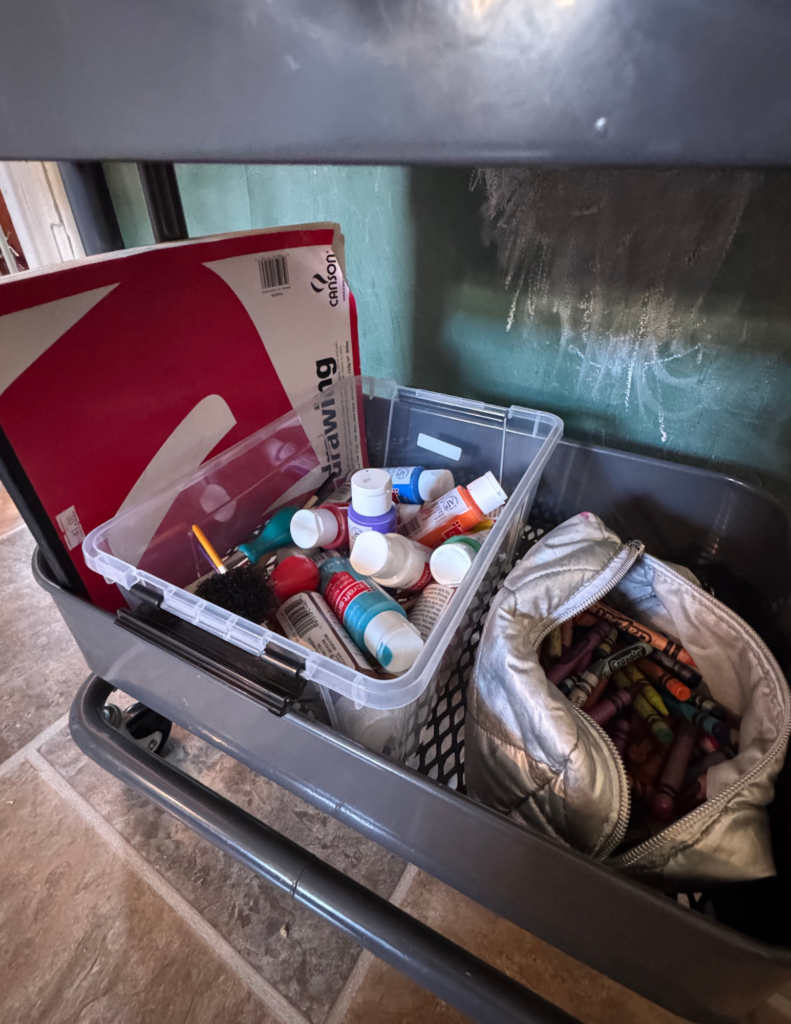 Bottom of rolling cart shelf with crayons in a pencil case, paints, and a paper pad
